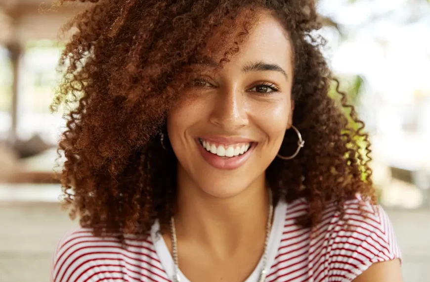 Uma mulher sorridente aparece em close, com cabelo cacheado castanho claro emoldurando o rosto. Ela usa uma camiseta listrada vermelha e branca e brincos de argola prateados. A expressão é alegre e confiante, transmitindo simpatia. O fundo é desfocado, sugerindo um ambiente ao ar livre ou com iluminação natural.