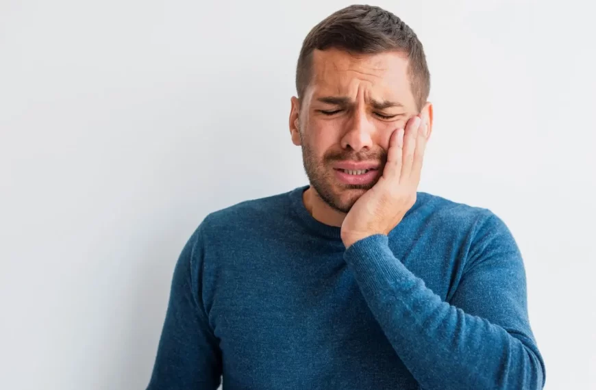 Um homem de cabelo castanho usando um suéter azul segura o rosto com a mão, exibindo expressão de dor e sofrimento; a cena sugere que ele quebrou um dente.