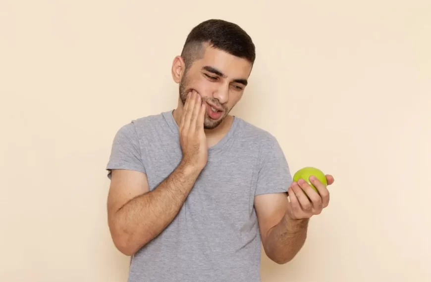 Jovem, com camisa cinza, segurando uma maçã verde e tocando a bochecha com expressão de dor — ilustração típica de desconforto imediato ao consumir alimento ácido ou crocante, sinalizando dentes sensíveis.