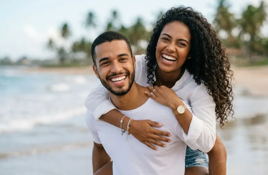 Um casal negro jovem e sorridente em uma praia tropical. O homem carrega a mulher nas costas (piggyback ride); ambos riem abertamente para a câmera. Ele veste uma camiseta branca simples, e ela usa uma camisa branca de botão e shorts jeans, com o oceano e coqueiros ao fundo sob um céu claro.